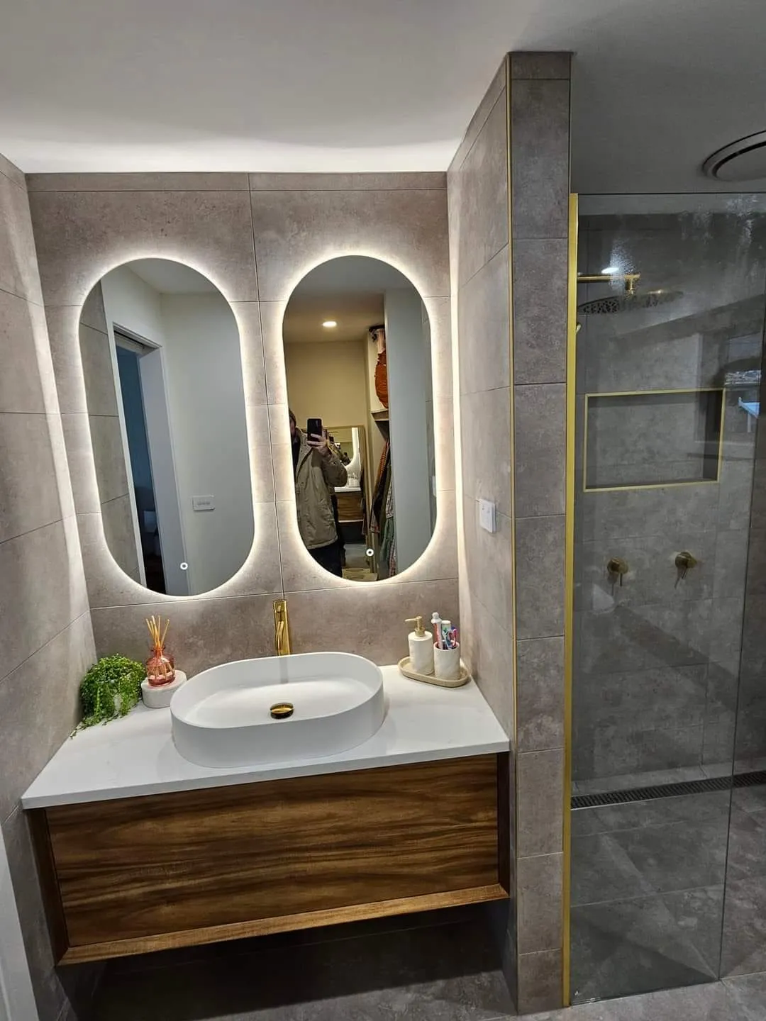 Timber vanity with a vessel basin and brass tap, below twin backlit oval mirrors