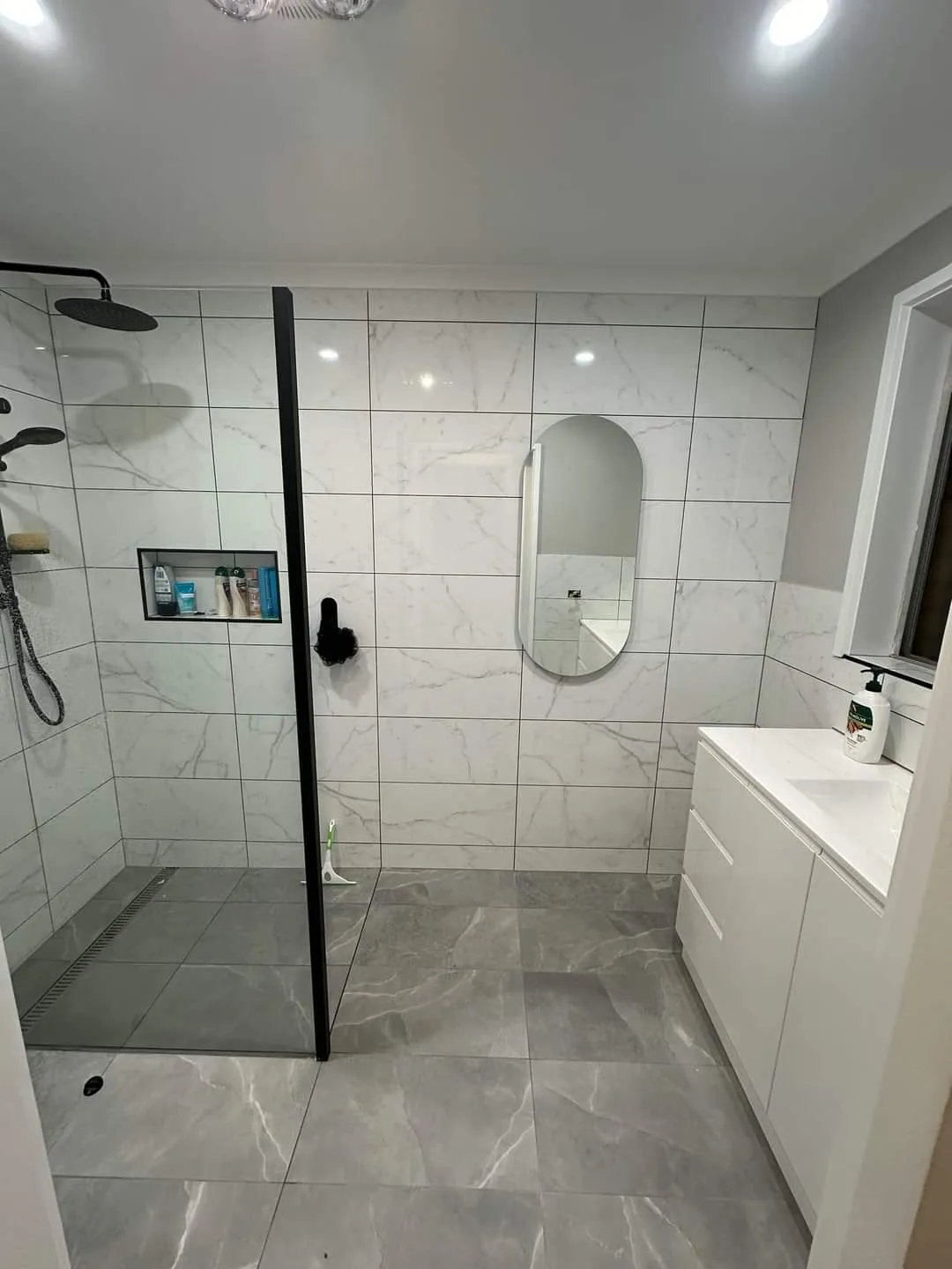 Marble-look tiled shower with black tapware and a black screen, beside a vanity and oval mirror