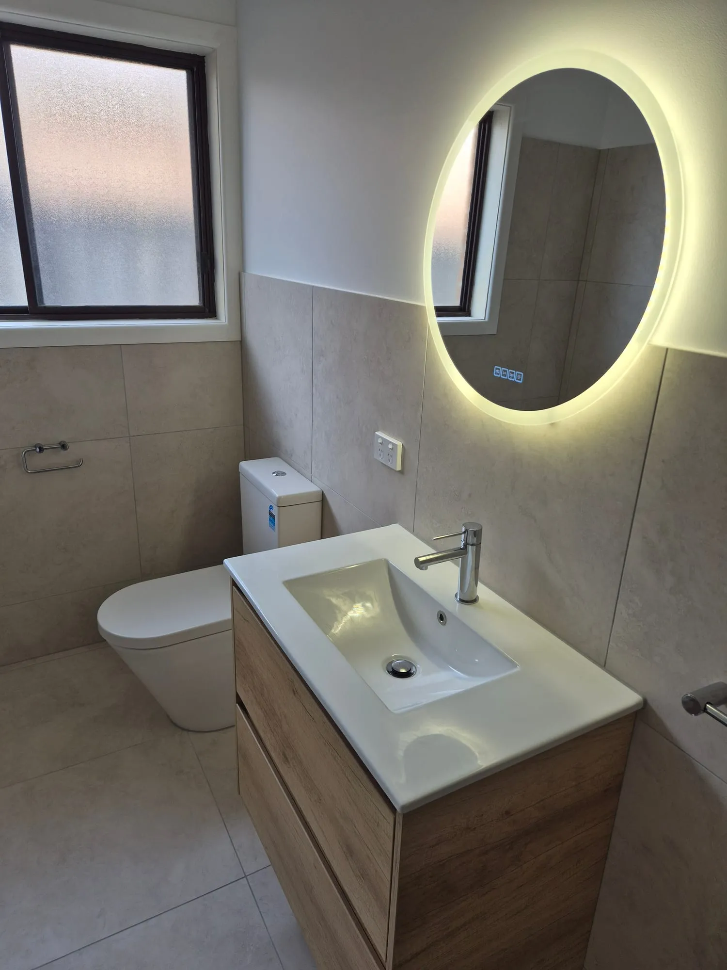 Timber vanity and toilet below a backlit round mirror