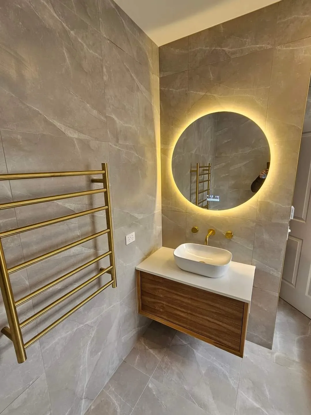 Timber vanity and vessel basin below a backlit round mirror, beside a brass towel ladder